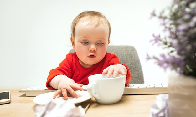 Glückliches Kindbaby, das mit Tasse und Tastatur des modernen Computers oder des Laptops lokalisiert auf Weiß sitzt