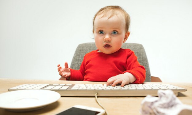 Glückliches Kindbaby, das mit Tastatur des modernen Computers oder des Laptops lokalisiert auf einem weißen Studio sitzt