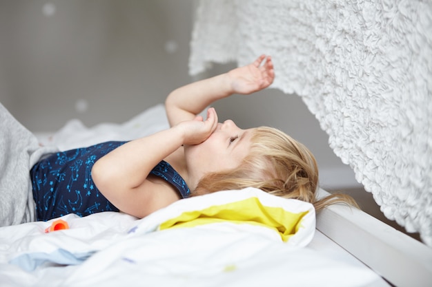 Glückliches Kindheitskonzept. Netter kleiner Junge mit blonden Haaren, die auf Bett in seinem weißen gemütlichen Schlafzimmer liegen und mit seinen Händen spielen.