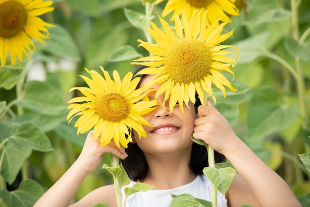 glückliches kleines asiatisches Mädchen, das Spaß unter blühenden Sonnenblumen unter den sanften Sonnenstrahlen hat.