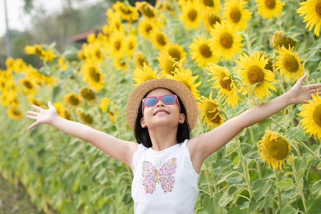 glückliches kleines asiatisches Mädchen, das Spaß unter blühenden Sonnenblumen unter den sanften Sonnenstrahlen hat.