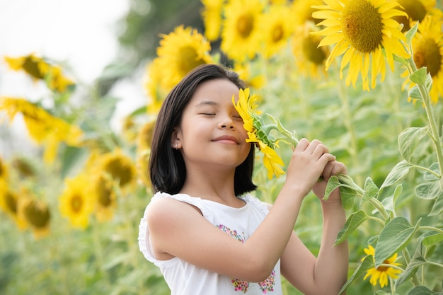 glückliches kleines asiatisches Mädchen, das Spaß unter blühenden Sonnenblumen unter den sanften Sonnenstrahlen hat.