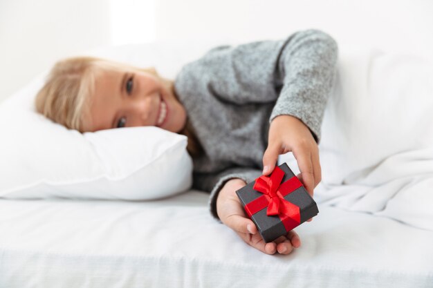 Glückliches kleines Mädchen, das kleine Geschenkbox hält, während im Bett liegend, selektiver Fokus