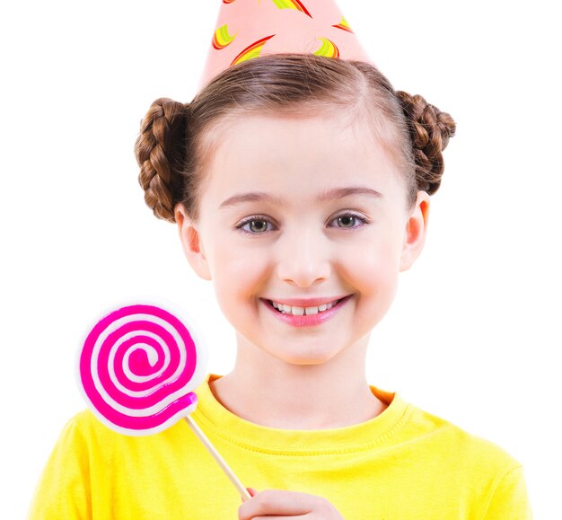 Glückliches kleines Mädchen im gelben T-Shirt und im Partyhut, die farbige Süßigkeiten halten - lokalisiert auf Weiß.