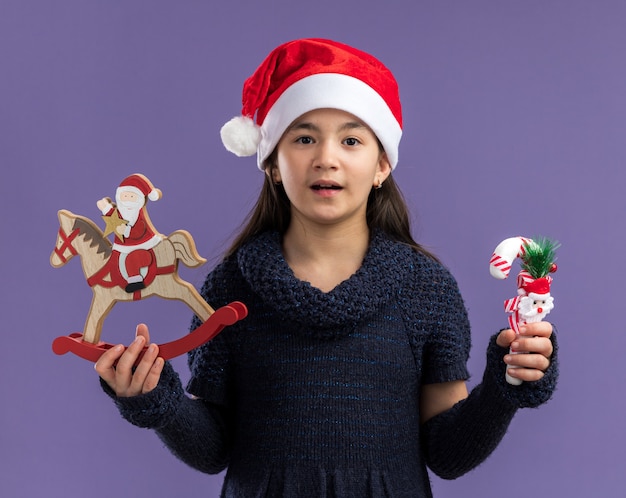 glückliches kleines Mädchen im Strickkleid, das Weihnachtsmütze hält, die Weihnachtsspielzeug hält, die Kamera betrachtet, die fröhlich über lila Hintergrund stehend lächelt