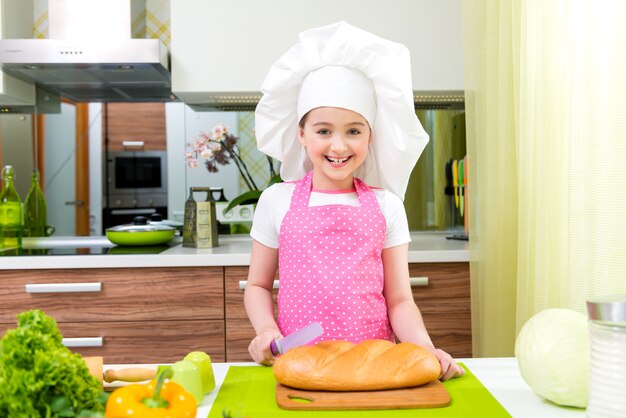 Glückliches kleines Mädchen in der rosa Schürze, die Brot in der Küche schneidet.