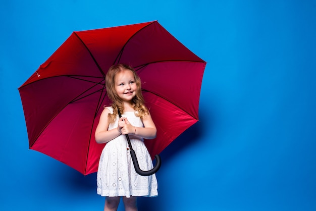 Glückliches kleines Mädchen mit rotem Regenschirm, der auf blauer Wand aufwirft.