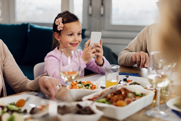 Glückliches kleines Mädchen mit Smartphone beim Mittagessen mit ihrer Familie zu Hause