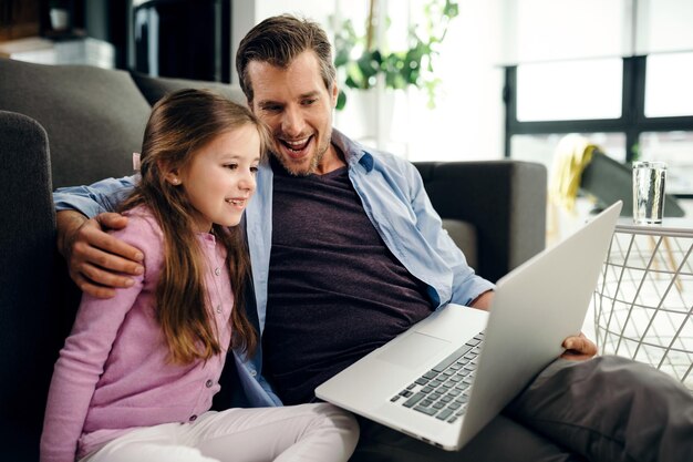 Glückliches kleines Mädchen und ihr Vater, die einen Computer benutzen und etwas im Internet ansehen, während sie sich im Wohnzimmer entspannen