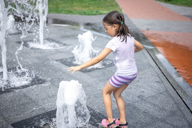 Glückliches kleines Mädchen unter dem Spritzwasser des Stadtbrunnens hat Spaß und entkommt der Hitze.