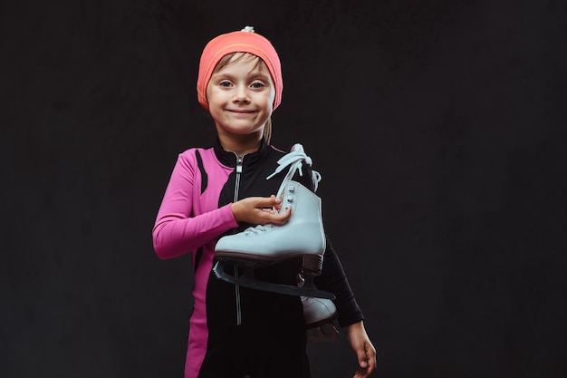 Glückliches kleines skatermädchen in sportbekleidung hält schlittschuhe auf einer schulter. Getrennt auf dunklem strukturiertem Hintergrund.