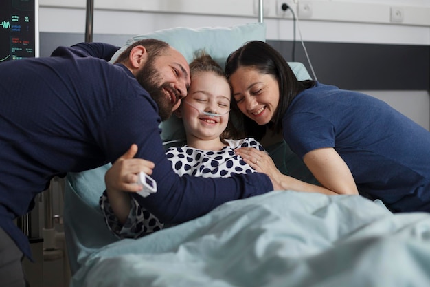 Glückliches krankes kleines Mädchen, das von fröhlich lächelnden Eltern in der Kinderstation des Krankenhauses umarmt wird. Fröhliche Mutter und Vater umarmen die kranke Tochter, die während der Behandlung im Patientenbett sitzt.