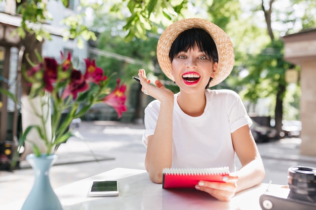 Glückliches lachendes Mädchen mit kurzen schwarzen Haaren hatte eine großartige Idee, mit Notizbuch und Stift im grünen Garten sitzend