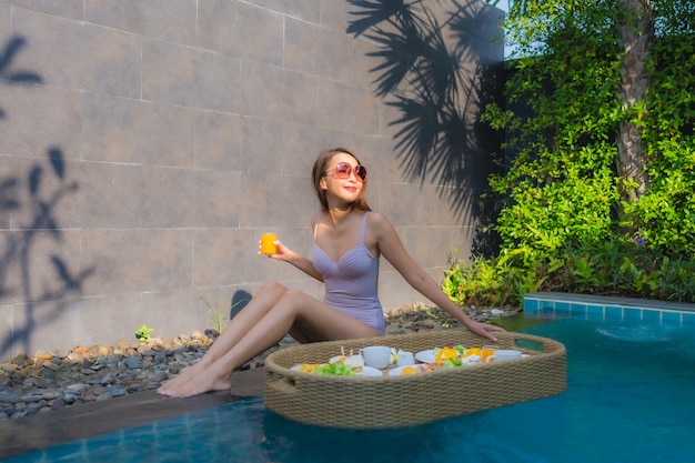 Glückliches Lächeln der jungen asiatischen Frau des Porträts genießen mit schwimmendem Frühstückstablett im Schwimmbad im Hotel