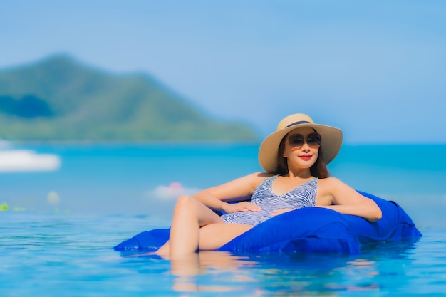 Glückliches Lächeln der schönen jungen asiatischen Frau des Porträts entspannen sich im Swimmingpool am neary Seeozeanstrand des Hotelerholungsortes auf blauem Himmel