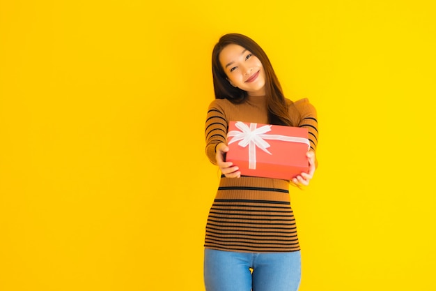 Glückliches Lächeln der schönen jungen asiatischen Frau des Porträts mit roter Geschenkbox auf gelber Wand