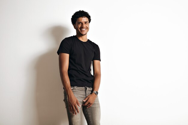 Glückliches lächelndes einladendes sexy junges schwarzes Männchen in engen grauen Jeans und leerem schwarzen Baumwoll-T-Shirt lokalisiert auf Weiß