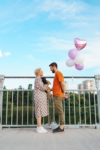 Glückliches lächelndes junges paar treffen auf brücke, die rosa luftballons und hund mit stadt am horizont hält