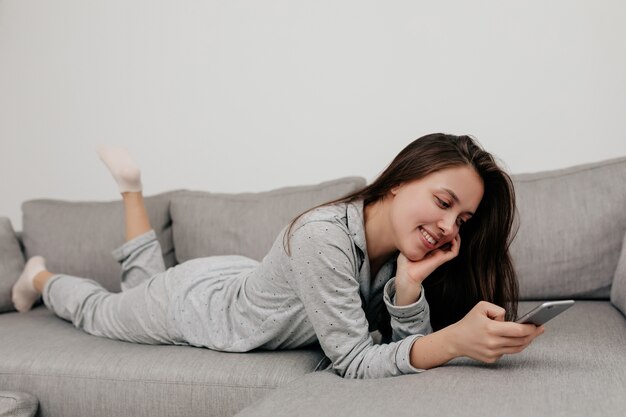 Glückliches lächelndes Mädchen in der Hauskleidung, die auf dem Trainer mit Smartphone ruht und ihre Massagen überprüft. Innenporträt zu Hause.