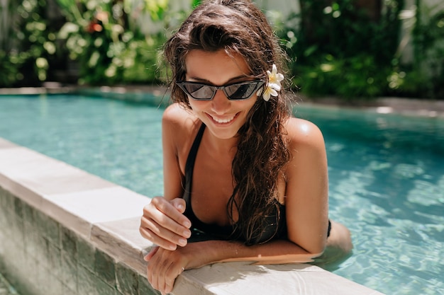 Glückliches lächelndes Mädchen mit dunklem nassem Haar im Badeanzug, der im Pool im sonnigen warmen Sommertag schwimmt. Hochwertiges Foto