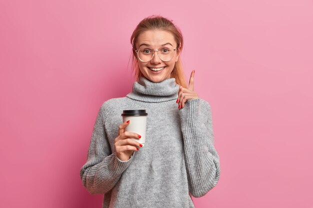 Glückliches lächelndes Mädchen mit positiven Ausdruckspunkten oben, hält heißes Getränk im Pappbecher, gekleidet im losen grauen Pullover