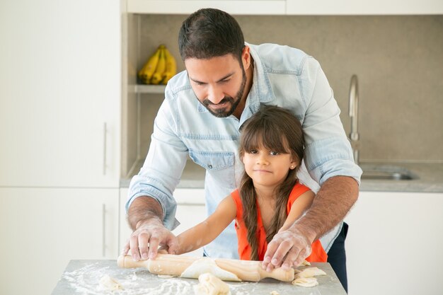 Glückliches lateinamerikanisches Mädchen und ihr Vater, die Teig auf Küchentisch mit Mehlpulver rollen und kneten.