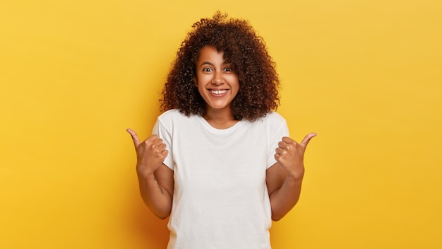 Glückliches lockiges Mädchen macht Daumen hoch Zeichen, zeigt Unterstützung und Respekt zu jemandem, lächelt angenehm, erreicht wünschenswertes Ziel, trägt weißes T-Shirt, isoliert auf gelber Wand