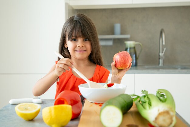 Glückliches Mädchen, das Apfel hält, während Salat in Schüssel mit großem Holzlöffel rührt. Nettes Kind, das lernt, Gemüse zum Abendessen zu kochen. Kochkonzept lernen