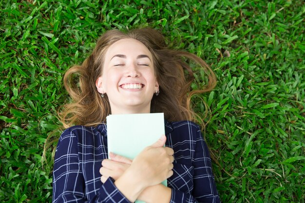 Glückliches Mädchen, das auf Gras und umarmendem Buch liegt