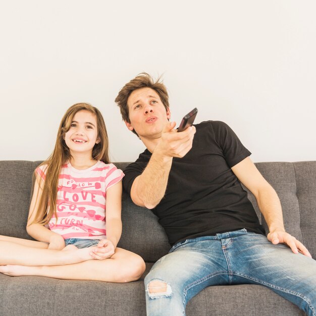Glückliches Mädchen, das auf Sofa mit ihrem Vater in der Hand halten Fernbedienung sitzt