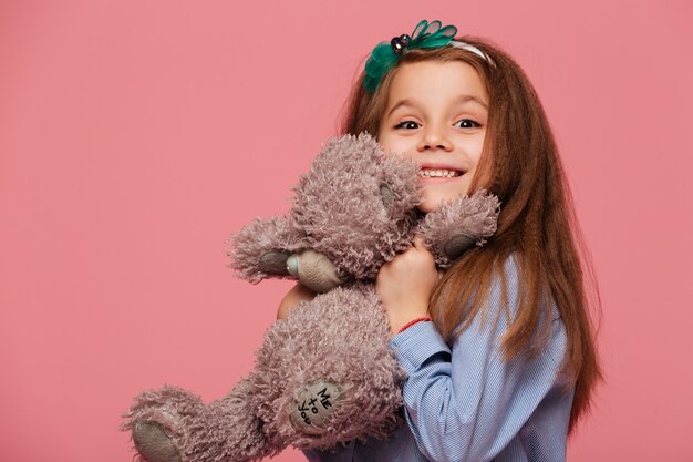 Glückliches Mädchen, das das lange kastanienbraune Haar lächelt hat, spielend mit ihrem reizenden Spielzeugteddybären