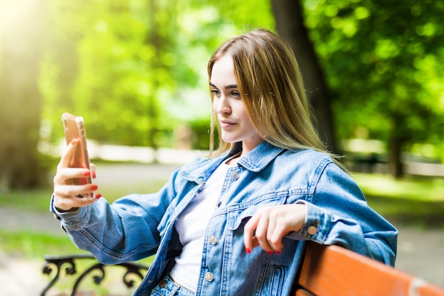 Glückliches Mädchen, das ein Telefon in einem Stadtpark benutzt, der auf einer Bank sitzt