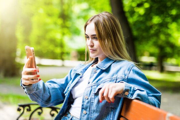 Glückliches Mädchen, das ein Telefon in einem Stadtpark benutzt, der auf einer Bank sitzt