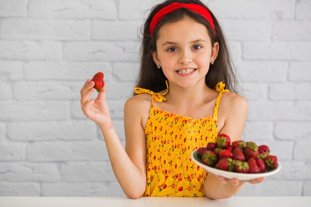 Glückliches Mädchen, das gegen die weiße Backsteinmauer hält rote Erdbeeren steht