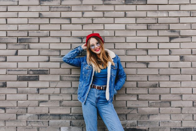 Glückliches Mädchen im Jeans-Outfit, das vor Ziegelmauer steht. Außenfoto der kaukasischen jungen Dame trägt Jeans und roten Hut, die positive Emotionen ausdrücken.