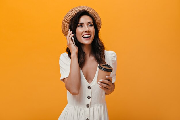 Glückliches Mädchen im Strohhut spricht am Telefon und hält Glas Kaffee. Schöne Dame in hellen Kleidern hält Smartphone und Tee auf lokalisiertem Hintergrund.