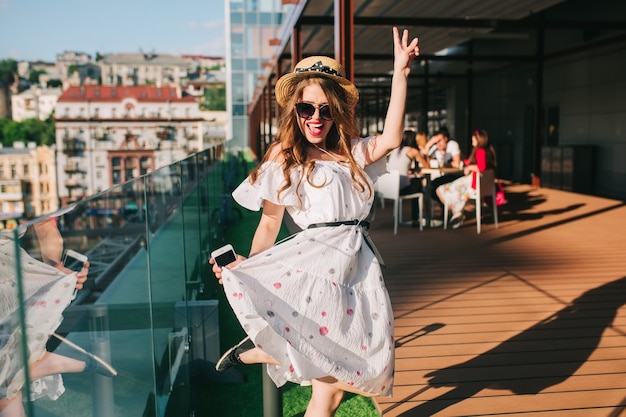 Glückliches Mädchen in der Sonnenbrille hört Musik durch Kopfhörer auf der Terrasse. Sie trägt ein weißes Kleid mit nackten Schultern, rotem Lippenstift und Hut. Sie tanzt.