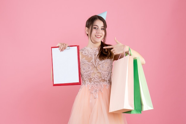 glückliches Mädchen mit der Partykappe, die Dokumente und Einkaufstaschen auf rosa hält