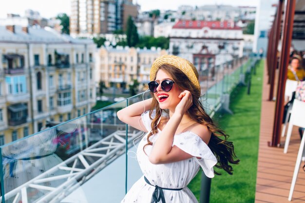 Glückliches Mädchen mit langen Haaren in der Sonnenbrille hört Musik durch Kopfhörer auf der Terrasse. Sie trägt ein weißes Kleid mit nackten Schultern, rotem Lippenstift und Hut.