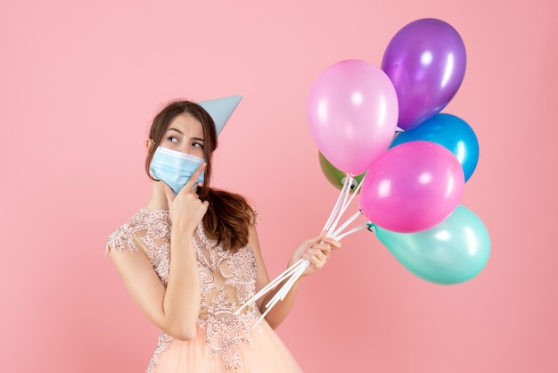 glückliches Mädchen mit Partykappe und medizinischer Maske, die bunte Luftballons hält, die Hand auf ihr Kinn auf Rosa setzen