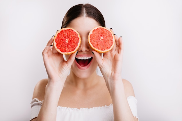 Glückliches Mädchen mit positivem emotionalem Gesichtsausdruck bedeckt ihre Augen mit Orangen, die auf isolierter Wand posieren.