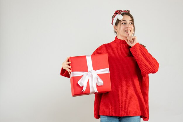 glückliches Mädchen mit Weihnachtsmütze, die Geschenk hält, das shh Zeichen auf Weiß macht