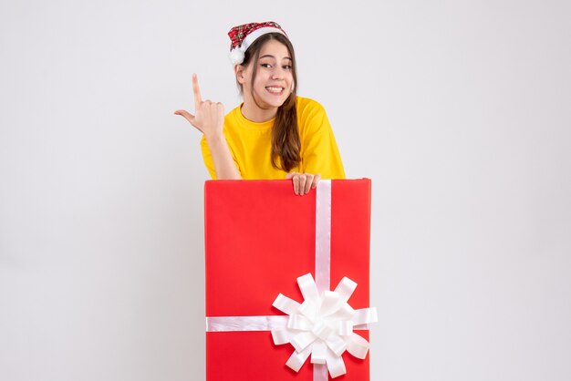 glückliches Mädchen mit Weihnachtsmütze, die hinter großem Weihnachtsgeschenk auf Weiß steht