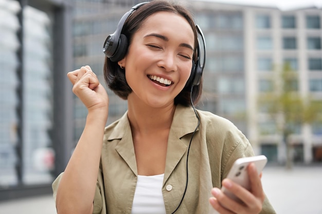 Glückliches Mädchen tanzt auf der Straße und hört Musik in drahtlosen Kopfhörern hält Handy mit Stream