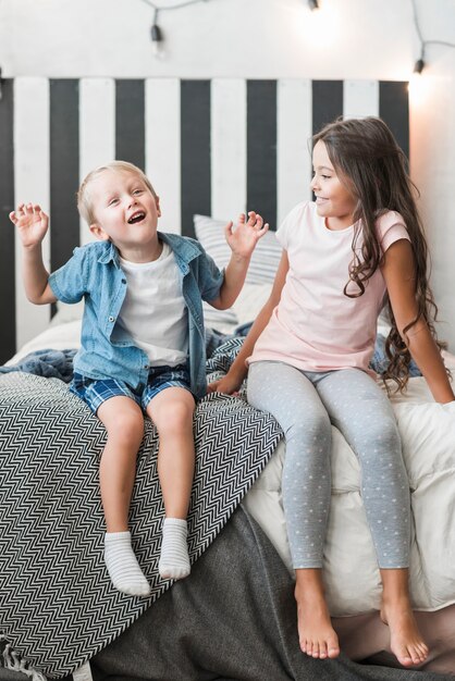 Glückliches Mädchen, welches den Jungen sitzt auf dem Bett tut Unfug betrachtet