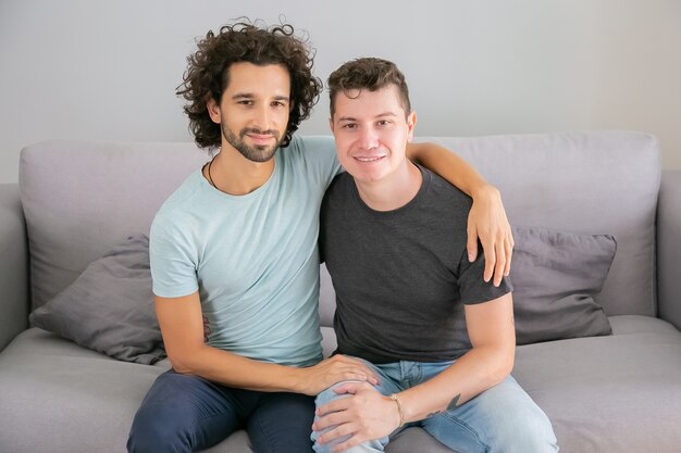 Glückliches männliches homosexuelles Paar, das zu Hause aufwirft, zusammen auf der Couch sitzt und sich umarmt. Vorderansicht. Liebes- und Beziehungskonzept