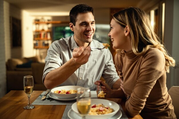 Glückliches Paar, das sich während einer Mahlzeit am Esstisch amüsiert Der Mensch füttert seine Freundin