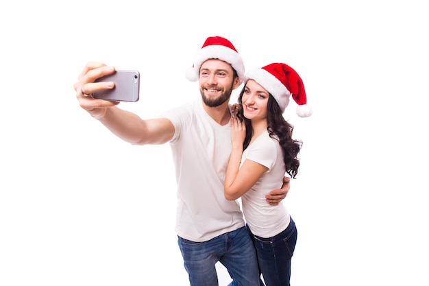 Glückliches paar in weihnachtsmützen winken und machen selfie auf dem smartphone, isoliert auf weißem hintergrund