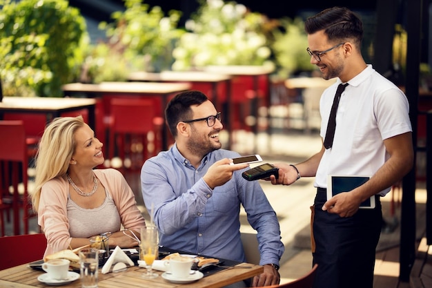 Glückliches Paar mit Handy und kontaktloser Zahlung an einen Kellner in einem Restaurant