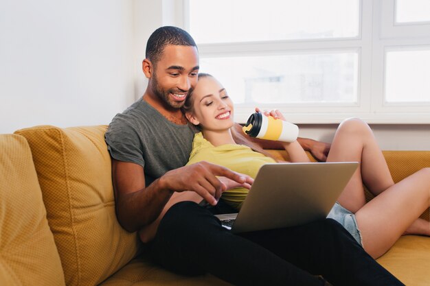 Glückliches Paar verbringt Zeit miteinander, kommuniziert, lächelt. Liebhaber umarmen und beobachten etwas auf Laptop, Mann und Frau sitzen auf Sofa